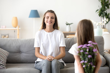 Obraz premium Cute little daughter hold flowers making surprise for mom greeting her with birthday, excited young mother sit on couch with eyes closed waiting getting gift from child, girl prepare present to mommy