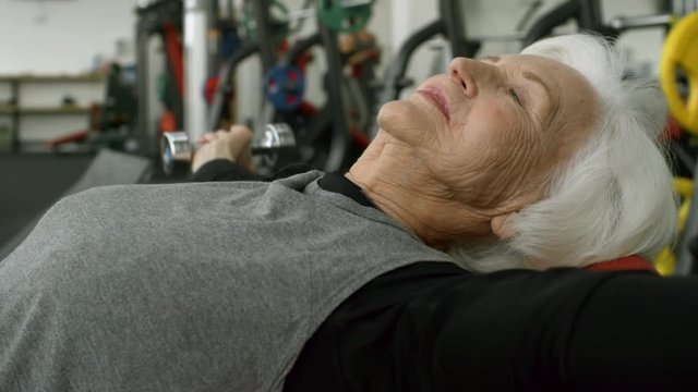 PAN of cheerful elderly woman lying on bench and training with dumbbells in gym