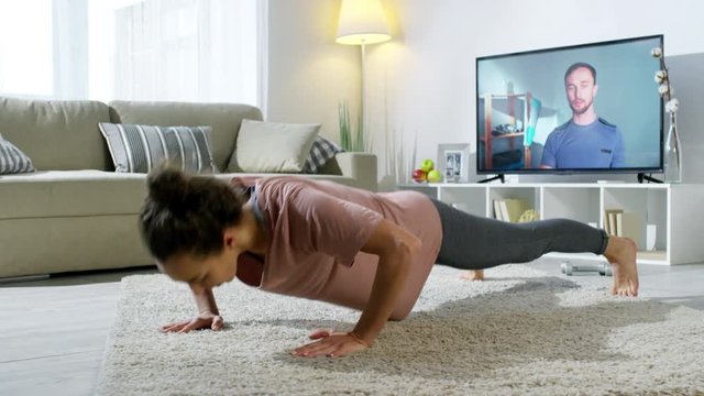 PAN of fit young woman in sportswear doing push-ups as her personal fitness trainer giving advice by video call on TV