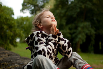 Little curly girl prays sitting on a stone fence. Rest in the village. Prayer to God. Summer...