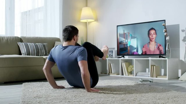 PAN of bearded man in sportswear doing knee tuck crunches on floor of his apartment and looking at TV screen as online personal fitness trainer instructing him by video call