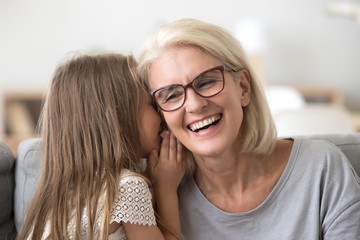 Cute small granddaughter whisper something in granny ear, grandmother and grandchild share secrets having sincere talk at home, grandma and little girl have fun at home together laughing - obrazy, fototapety, plakaty