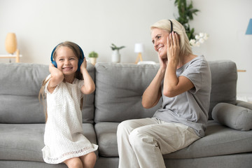 Excited grandmother and smiling little granddaughter listen to music in headphone, grandma and grandchild have fun relaxing on couch at home together, granny enjoy spending time with young generation