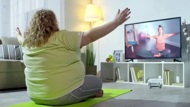 PAN of overweight woman sitting in lotus pose and stretching arms with online personal yoga instructor on TV screen