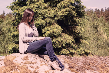 Naklejka na ściany i meble Young pretty girl is talking on the phone while sitting on a large stone against the background of a big fir