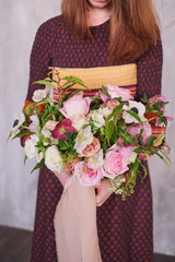 Young female florist holding a freshly made blooming floral bouquet of pastel pink carnations and eucalyptus against a gray wall.