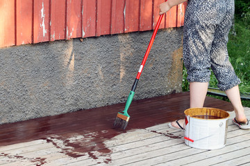 Woman oiling a wooden terrace with wood oil . She is using a paintbrush with pole extension. Copy space.