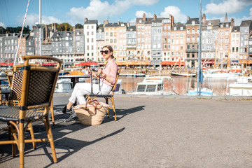 Young woman enjoying coffee sitting at the cafe outdoors near the harbour with beautiful buildings on the background in Honfleur old town, France
