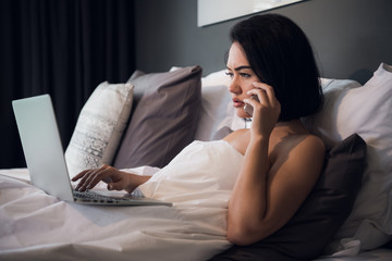 Smiling relaxed young woman using mobile phone and laptop in bed at home