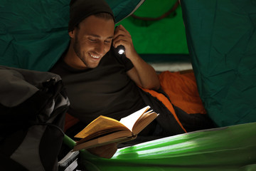 Young man in sleeping bag reading book with flashlight inside of tent