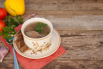 Clear beef broth, bouillon in white bowl and vegetables on wooden table. Rustic style.