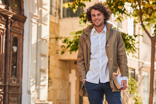 Outdoor image of handsome young man with books outdoors. College male student carrying books in college campus in autumn street background. Smiling cheerful guy with curly hair posing with books.