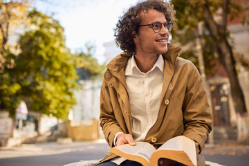 Happy young man with glasses reading and posing with book outdoors. College male student carrying books in campus in autumn street. Smiling guy wears spectacles and curly hair reading books outside