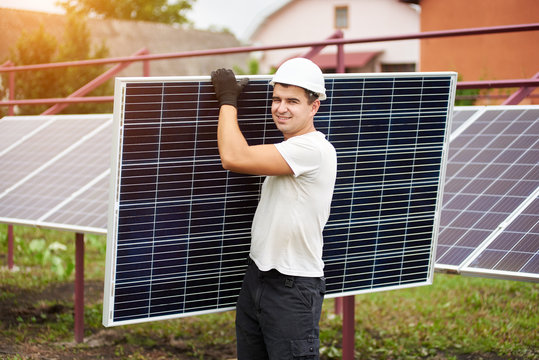 Smiling young worker in protective helmet carrying big shiny solar photo voltaic panel on exterior metal platform background on sunny summer warm day. Renewable ecological green energy concept.