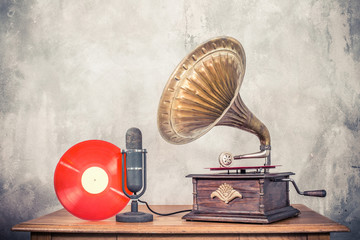 Vintage antique gramophone phonograph turntable with brass horn, studio microphone and red color vinyl discs on wooden table front concrete wall background. Retro old style filtered photo