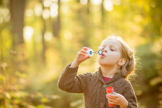 Portrait of funny lovely little girl blowing soap bubbles.Cute blonde blue-eyed girl in yellow knitted coat in the autumn park has fun. Happy funny childhood concept..Copy space