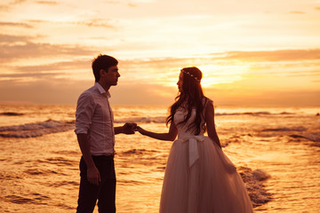 A beautiful couple of newlyweds, the bride and groom walking on the beach. Gorgeous sunset and sky. Wedding dresses, a white luxury dress for a girl. Family concept, honeymoon.