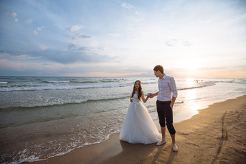 A beautiful couple of newlyweds, the bride and groom walking on the beach. Gorgeous sunset and sky. Wedding dresses, a white luxury dress for a girl. Family concept, honeymoon.
