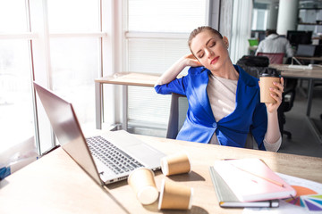 Hard-working woman is sitting at the table and has some rest. She is bored and tired. Woman has a cup of coffee in her hands. Also she is stretching with her eyes closed.