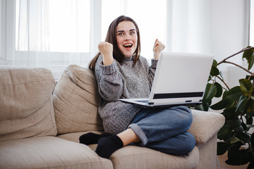 Happy woman winning online. Young excited lady using laptop in living room. Achievement, freelance, success concept