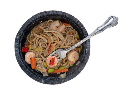 Top view of a shrimp and noodles with vegetables in a ginger sauce TV dinner in a black paper tray with a fork in the food isolated on a white background.