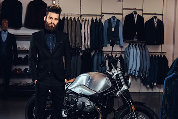 Fototapeta na wymiar Bearded stylish man dressed in black suit posing with hands in pockets near retro sports motorbike at the men's clothing store.