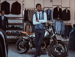 Elegantly dressed African American young man posing with crossed arms near retro sports motorbike at the men's clothing store.