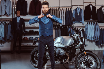 Stylish man dressed in elegant wear posing with hands on bow tie near retro sports motorbike at the men's clothing store.