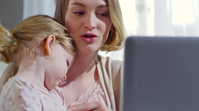 Tilt up of beautiful young mother holding shy toddler girl on her laps and talking on video call on laptop