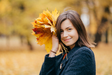 Cute smiling young European woman has stroll in park, enjoys sunny day during autumn, carries yellow leaves, wears coat, poses against blurred background. People, leisure and season concept.