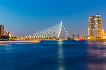 Cityscape on a calm night with illuminated Erasmus cable-stayed bridge over the river Maas by buildings against clear blue sky, yellow lights, Rotterdam in the Netherlands. Long exposure photography