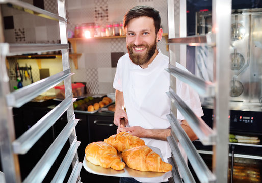 Handsome baker in white uniform holding in his hands a tray full of freshly baked croissants against the background of a bakery