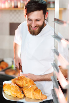 A young handsome male baker holds a tray with French croissants in front of a bakery and smiles.