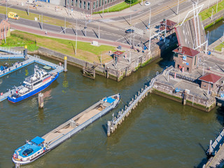Fototapeta na wymiar Aerial view of a cargo ship sailing on New Maas river towards a canal with raised drawbridge, boat anchored at pier, vehicular street in background, sunny day in Rotterdam, South Holland, Netherlands