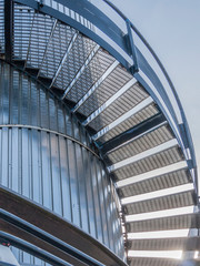 Fototapeta premium Low angle perspective of exterior metal spiral or elliptical stairs with railing against a sheet metal wall in a tower, grating tread or galvanized steel grille, sunny day with a blue sky