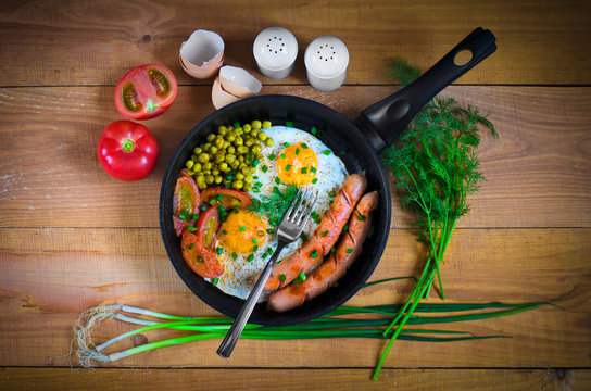 scrambled eggs in a frying pan with sausages and vegetables on wooden background