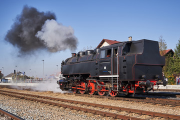 Retro steam train locomotive in the vilage railway station making smoke and steam and waiting on the track to be connected with railway carriage during the sunny day.