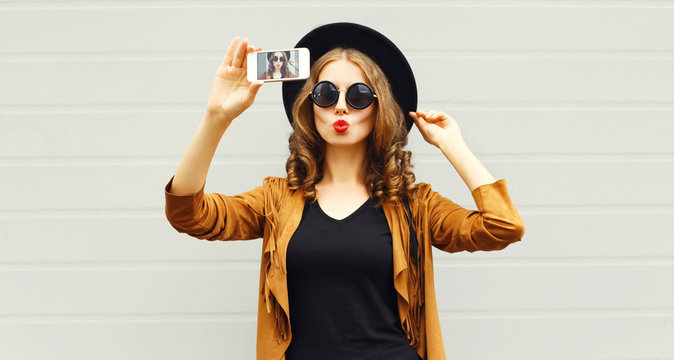 Fashion woman taking picture selfie by smartphone in black round hat, sunglasses, brown jacket on gray wall background
