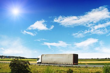Fototapeta na wymiar Truck with the trailer on the countryside road with fields and trees against blue sky with clouds and sun