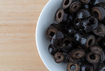 Top close view of a white bowl filled with sliced olives on a wood table.