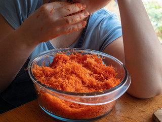 Young girl in apron and a cook's cap prepares fresh carrot salad in kitchen. Woman cook rubs carrots through grater, adds garlic, salt and mayonnaise