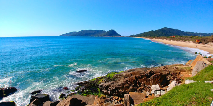 Caldeir&atilde;o and Arma&ccedil;&atilde;o beaches in Florianopolis, Brazil, during a sunny morning. Rocks can be seen close to the photographer, and mountains can be seen far away.