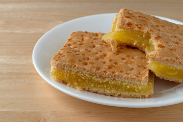 Close side view of two freshly baked lemon squares with one bitten on a white plate atop a wood table.