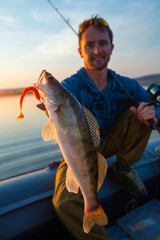 Young angler holds fish (zander) sitting in a boat at a lake during sunset