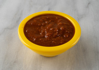Side view of hot dog chili sauce in a bright yellow bowl atop a gray marble counter top.
