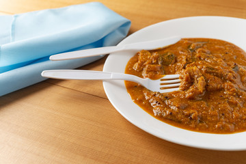 A meal of eggplant curry on a white plate with a plastic fork and knife in the food plus a blue napkin to the side atop a wood table top.