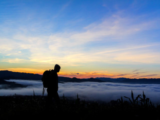 Silhouette of tourist man standing on top mountain and looks into the distance at morning sunrise with foggy mountain landscape