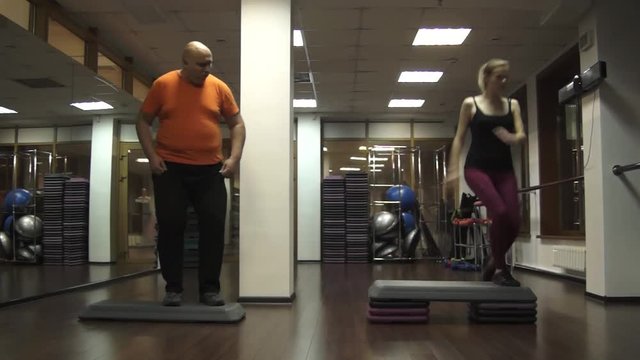 Fat funny man trying to do steps. Near young beautiful young sport woman actively doing step aerobics