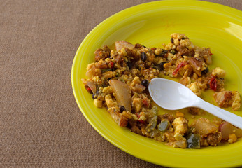 A partially eaten scrambled burrito meal on a yellow plate with a white spoon atop a brown tablecloth.