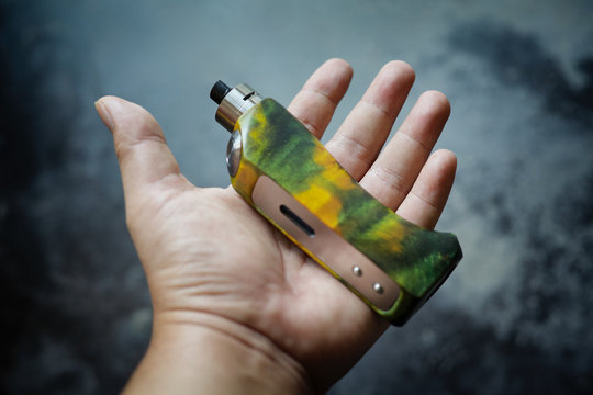 high end yellow green stabilized wood regulated box mods with rebuildable dripping atomizer in hand on dark grey texture background, vaping device, selective focus